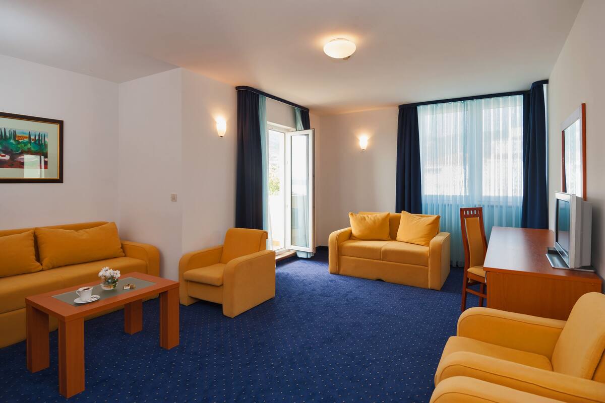 A hotel room with a blue carpet, yellow sofas, a wooden coffee table, and a TV on a wooden stand.