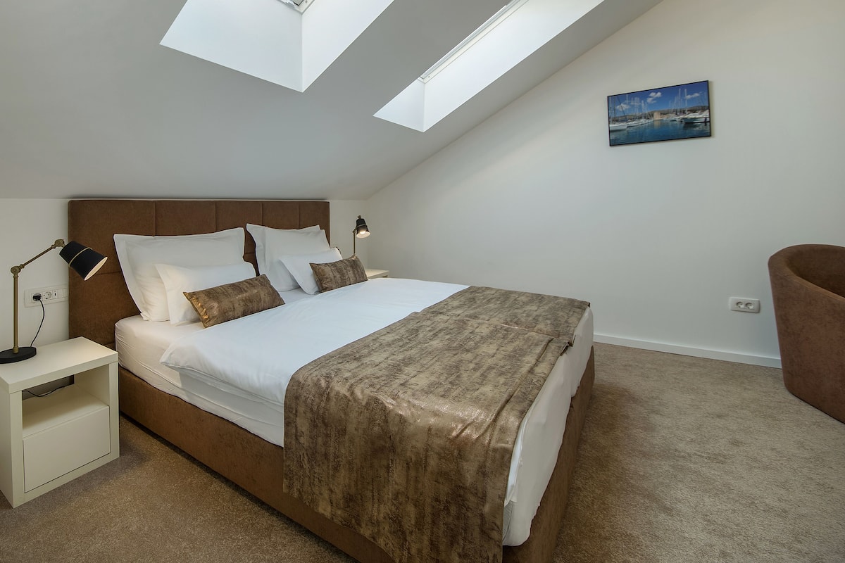 A bedroom with a bed, bedside tables, a nightstand, a lamp, a painting on the wall, and a skylight.