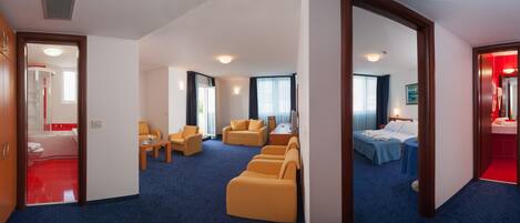 Quarto de hotel com carpete azul, poltronas amarelas, cama com colcha azul e banheiro vermelho visível por uma porta aberta.