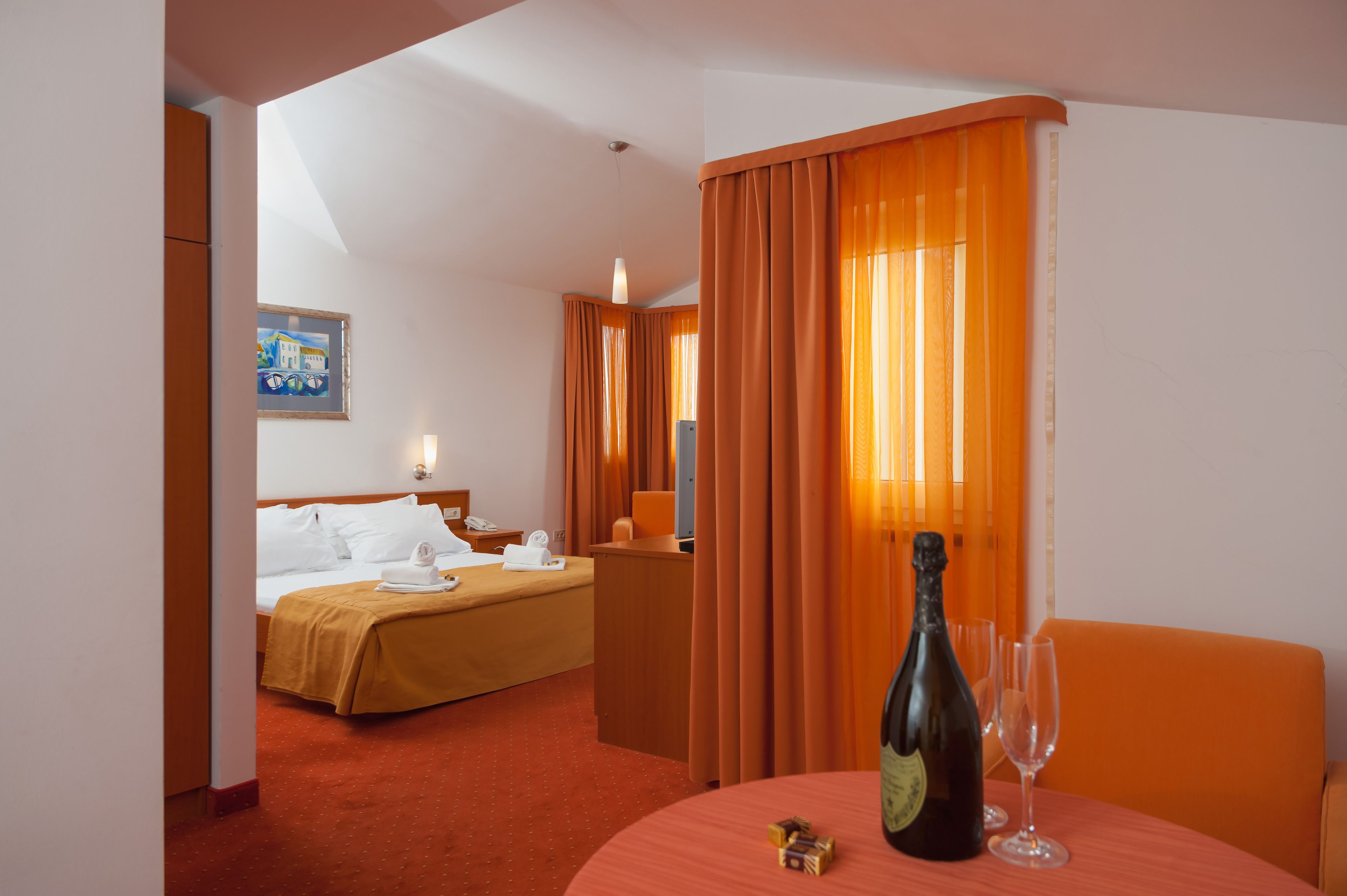 A hotel room with a bed, a desk, a table with a bottle and glasses, and orange curtains.