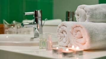 Une salle de bain avec un lavabo blanc, deux serviettes enroulées, un verre d’eau et trois bougies allumées.