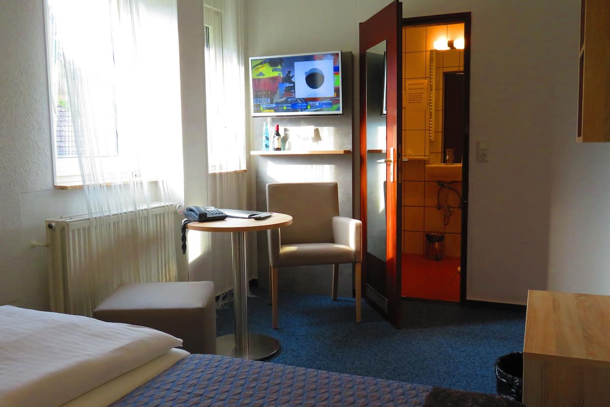 Une chambre d’hôtel avec un lit, une petite table et une chaise, un téléviseur fixé au mur, et une salle de bain visible à travers une porte ouverte.