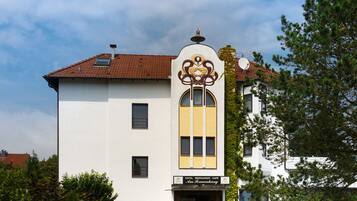 A two-story building with a white facade and a red-tiled roof. It features a central entrance with a decorative archway and a sign indicating it is a hotel and restaurant.