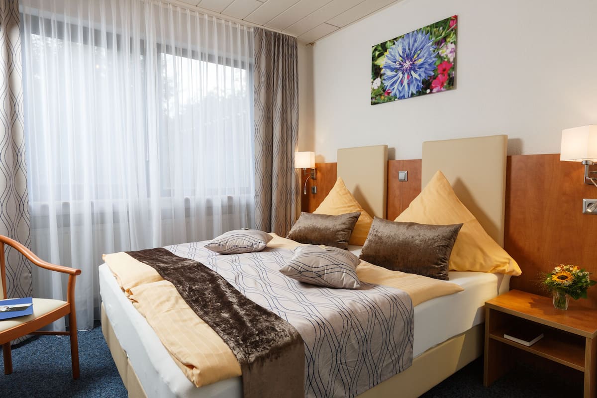 Une chambre d’hôtel avec un grand lit, deux tables de chevet, une chaise, une fenêtre avec des rideaux légers et un tableau représentant des fleurs accroché au mur.