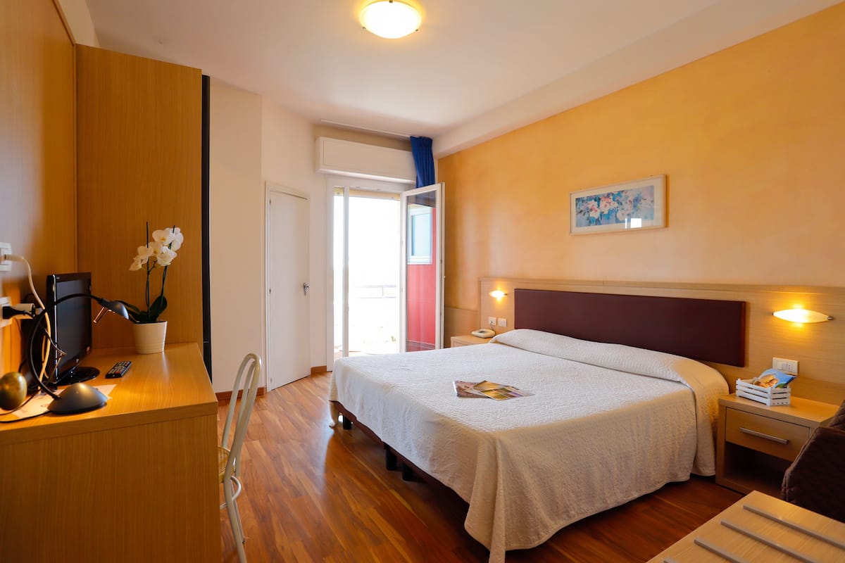 Ein Hotelzimmer mit Bett, Nachttischen, einem Schreibtisch mit Computer und Pflanze sowie einer Tür zu einem Balkon.