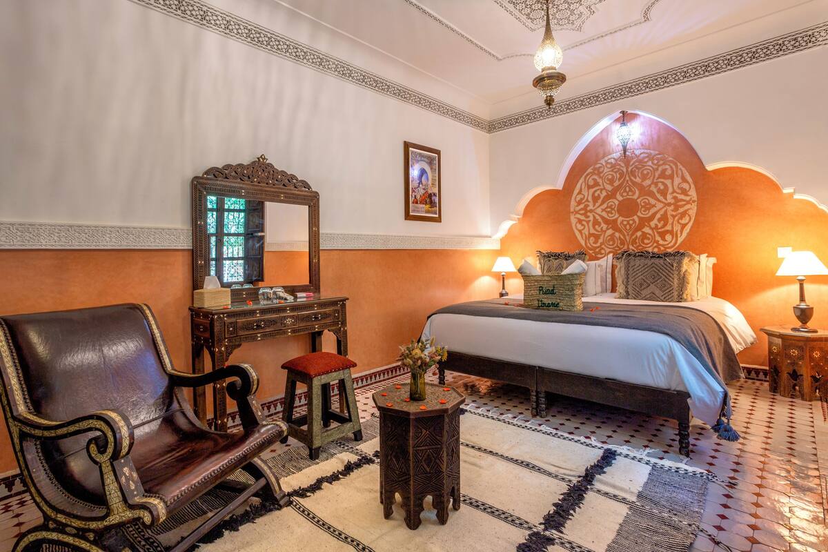 Habitación de hotel con cama, una silla, una mesita con un florero, un espejo y un adorno en la pared.