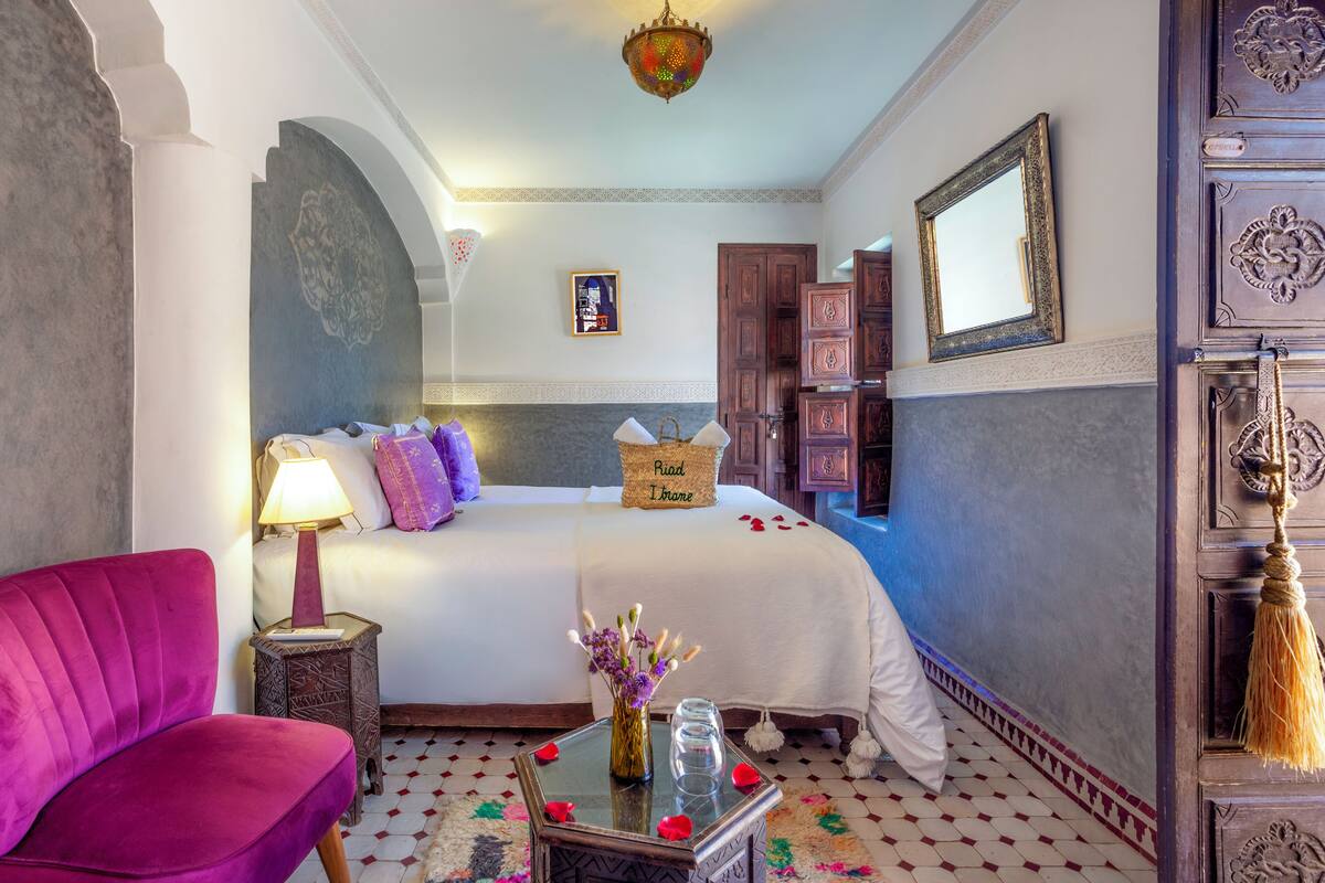 Una habitación de hotel con una cama, una silla, una mesa con un jarrón de flores y un espejo en la pared.
