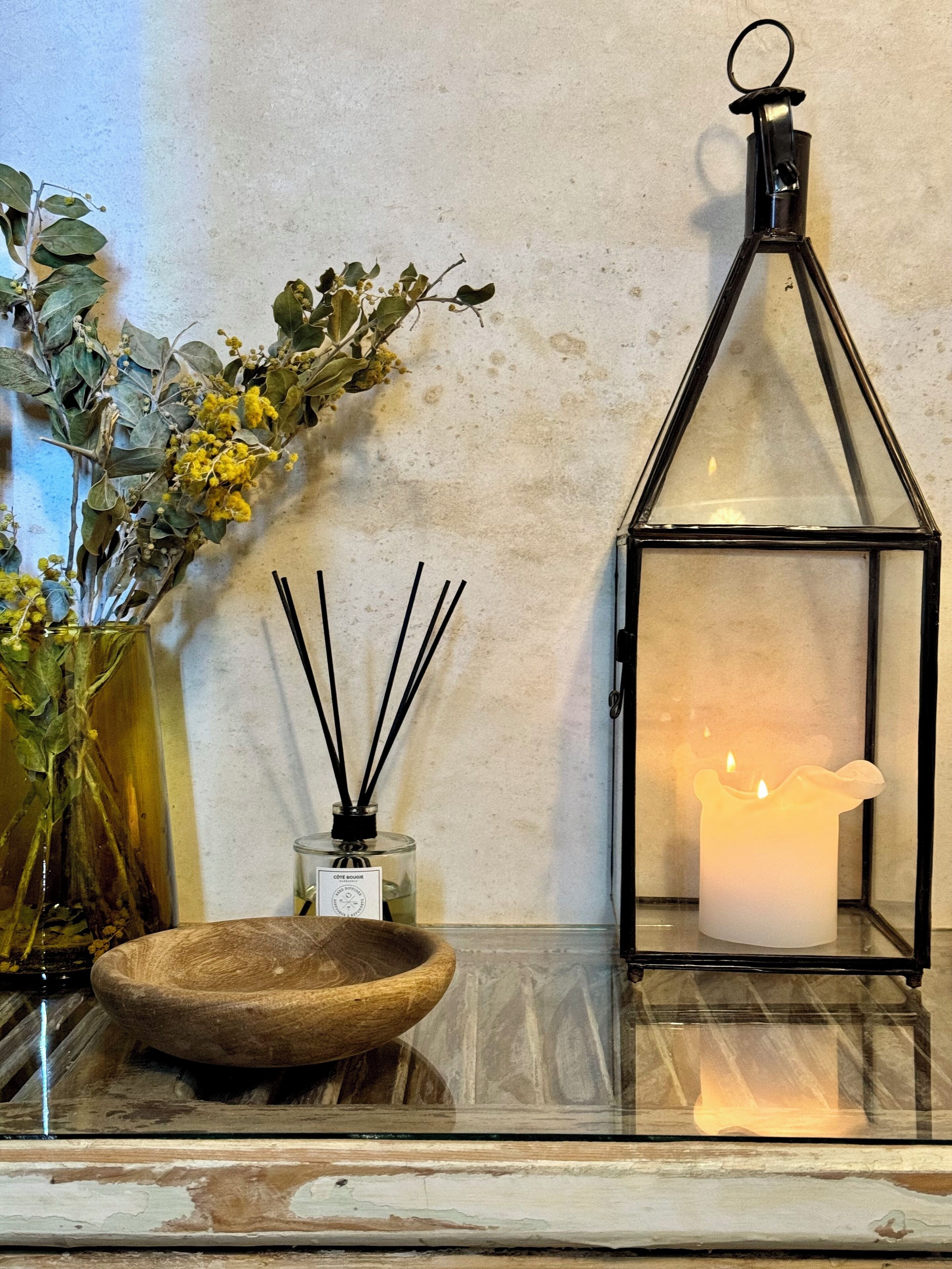 A candle in a glass lantern, a small bowl, and a vase with dried flowers and greenery.