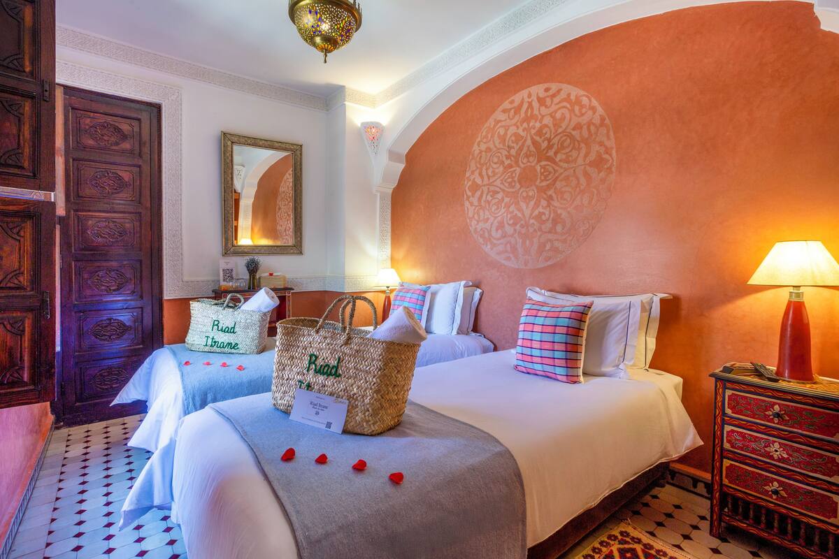 Habitación de hotel con dos camas, un espejo grande, un adorno en la pared, una puerta de madera y una mesita de noche con lámpara.