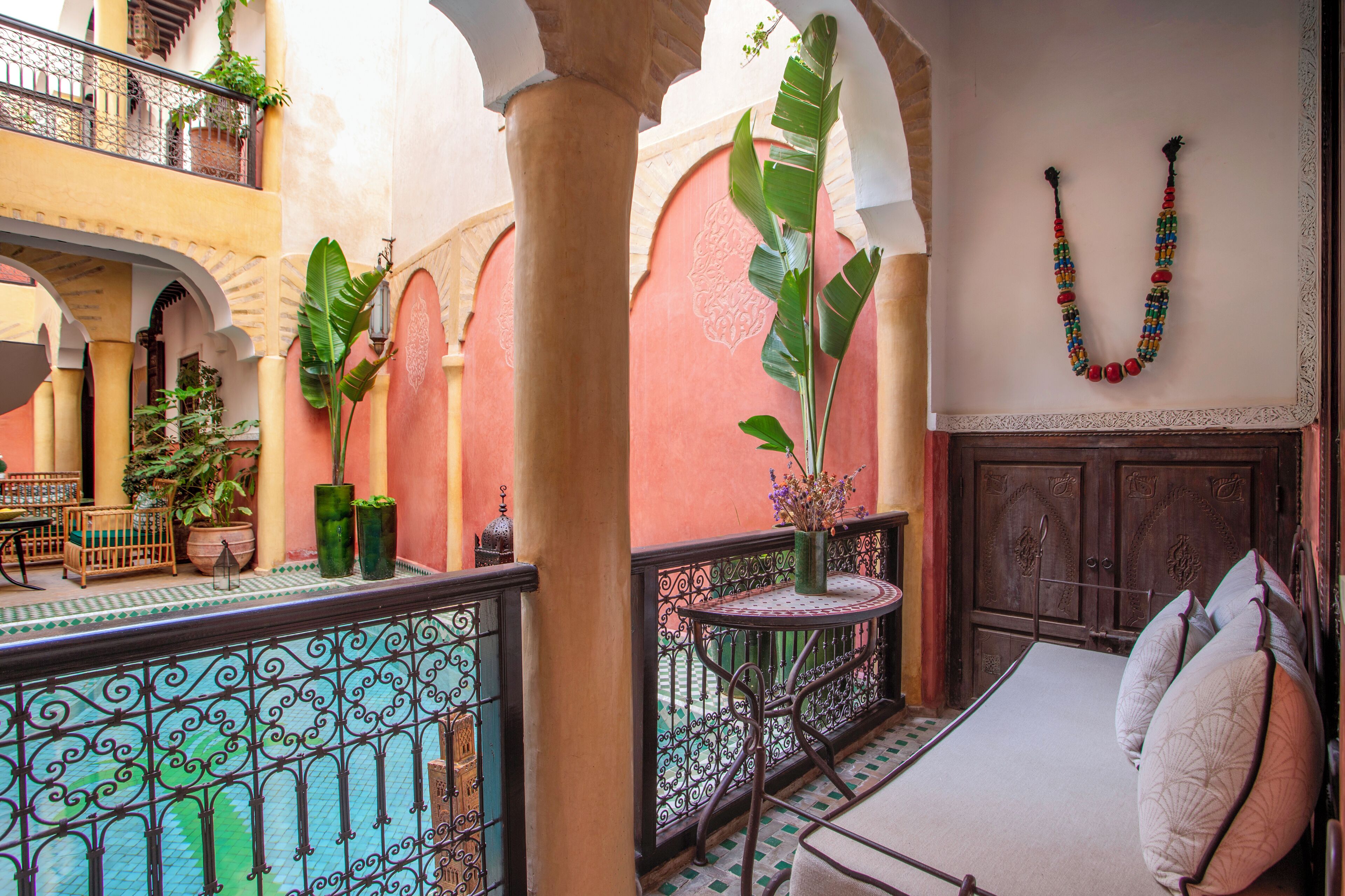 A traditional Moroccan interior with a balcony, a bed with pillows, a decorative wall hanging, and potted plants.