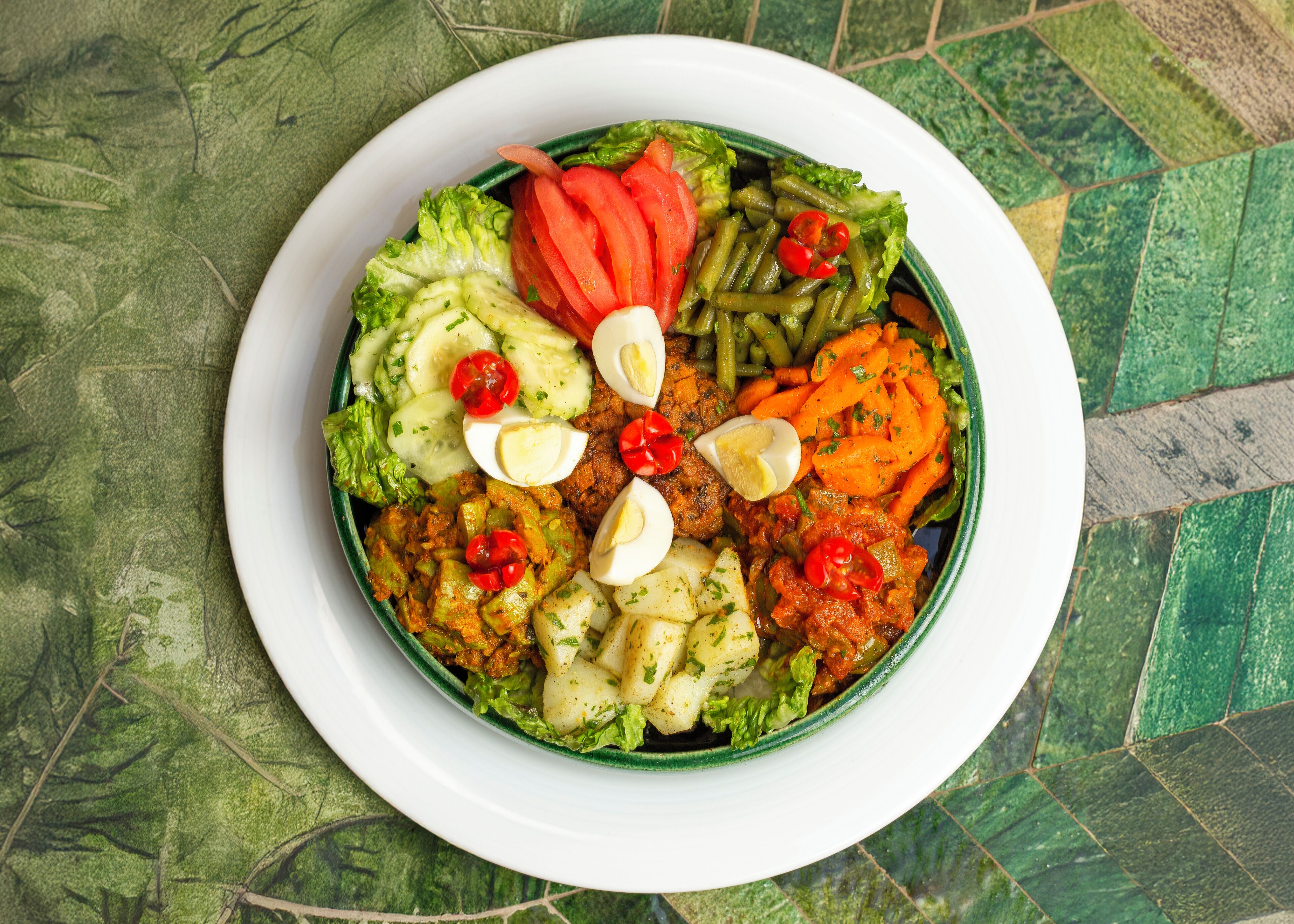 A bowl of salad with various vegetables and eggs on a tiled table.
