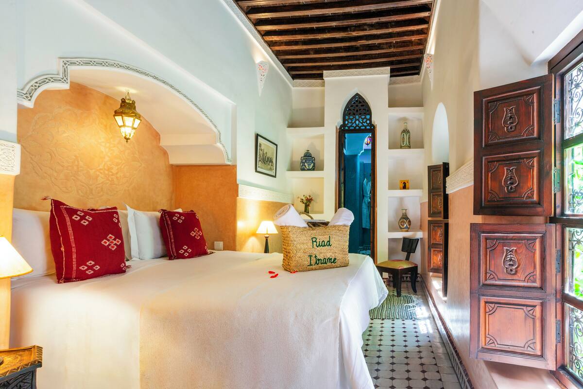 Habitación de hotel con una cama grande, una lámpara de noche, una cesta de mimbre con la etiqueta 'Fiad Ifrane' y vista al pasillo.