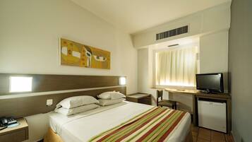 Quarto de hotel com cama, escrivaninha com televisão, pequeno frigorífico, mesa de cabeceira e um quadro na parede.