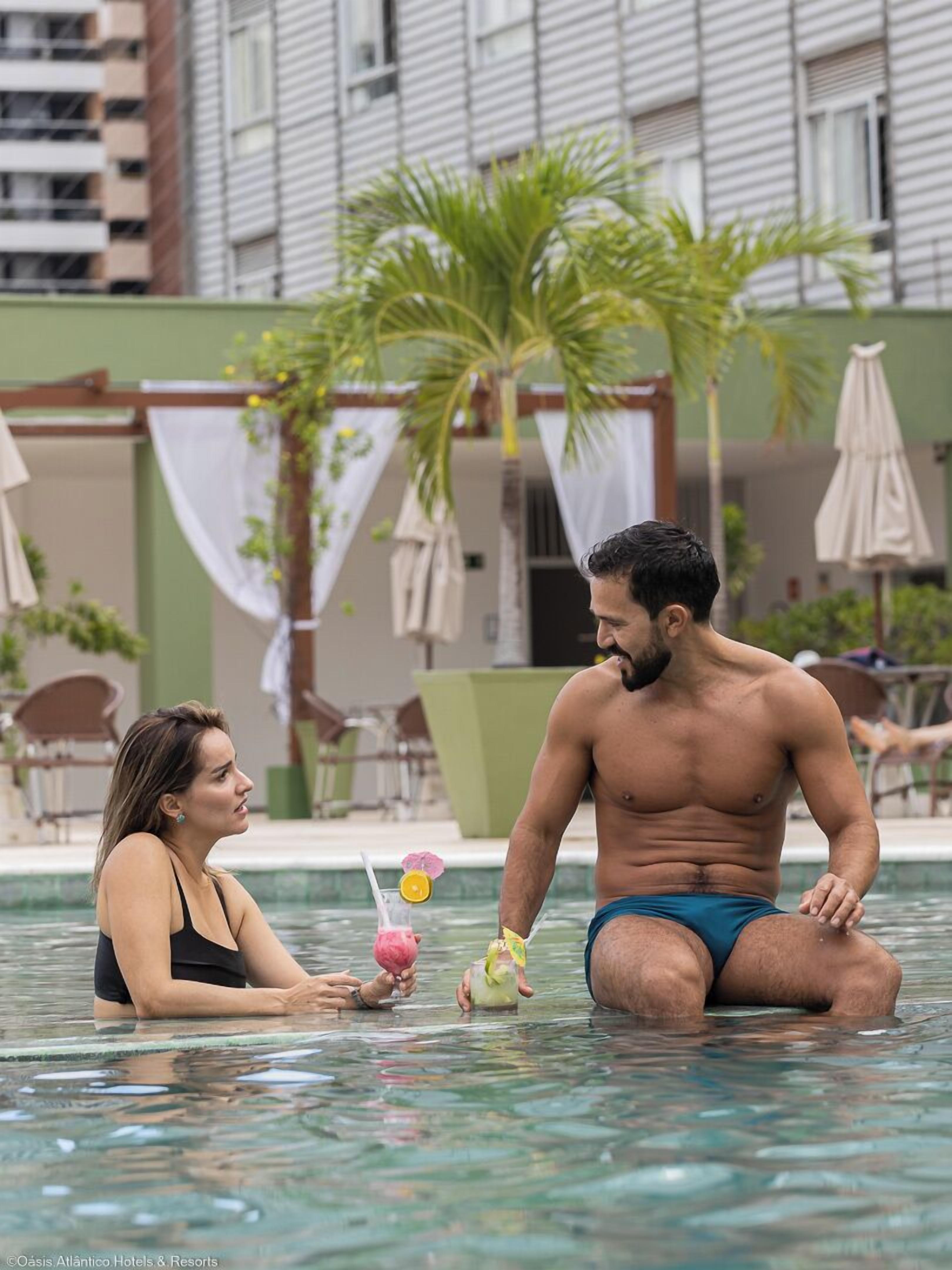 Un hombre y una mujer en una piscina, con un entorno tropical que incluye palmeras y un edificio al fondo.