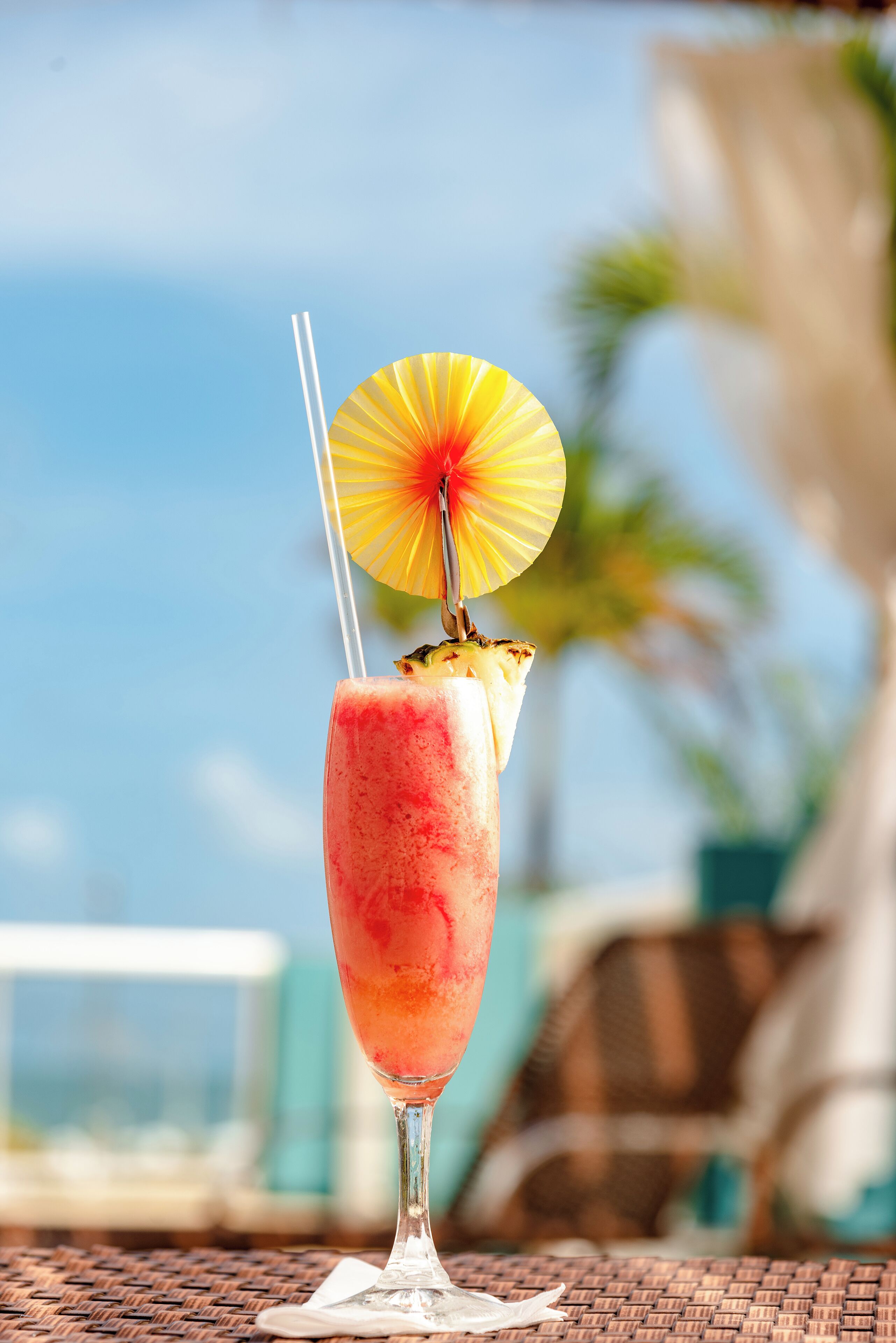A tropical drink with a straw and a pineapple slice on a table with a wicker chair and a view of the ocean.