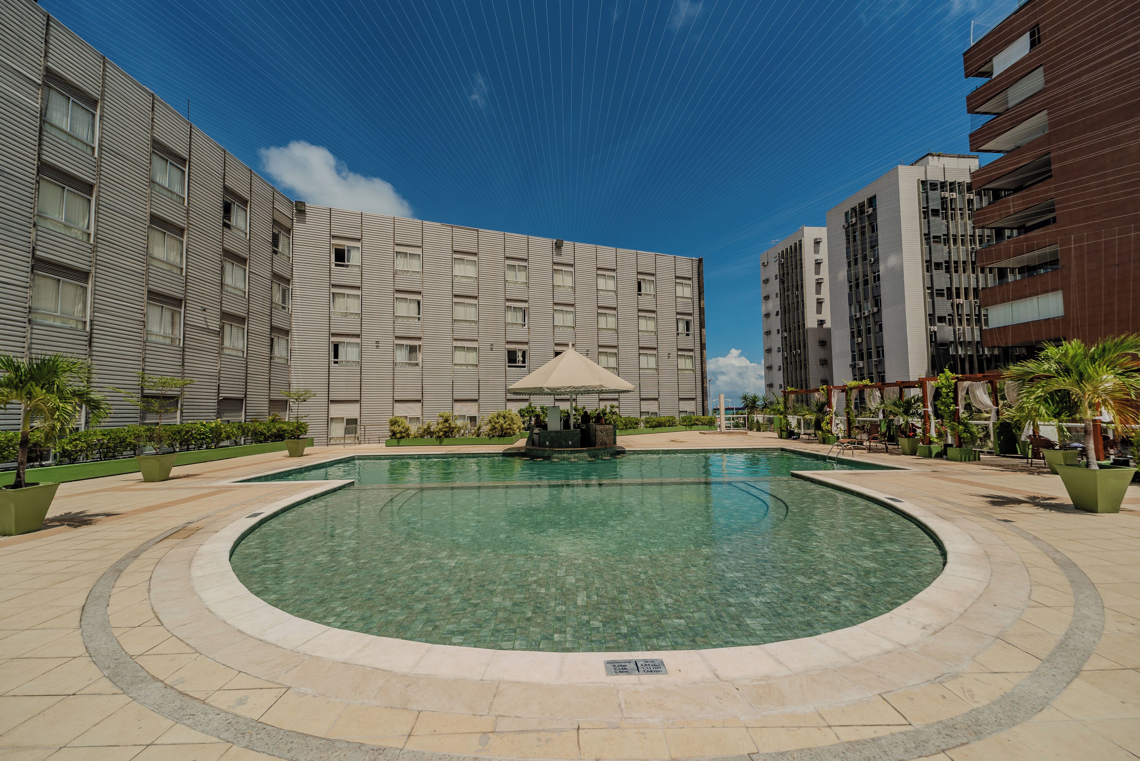 A modern hotel courtyard with a central swimming pool, surrounded by greenery and adjacent to multi-story buildings.
