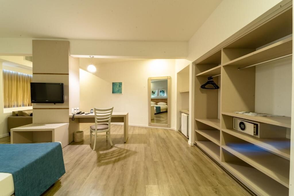 Quarto de hotel com cama, escrivaninha, cadeira, televisão, guarda-roupa e espelho.