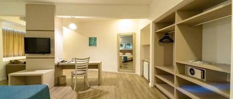 Quarto de hotel com cama, escrivaninha, cadeira, televisão, guarda-roupa e espelho.