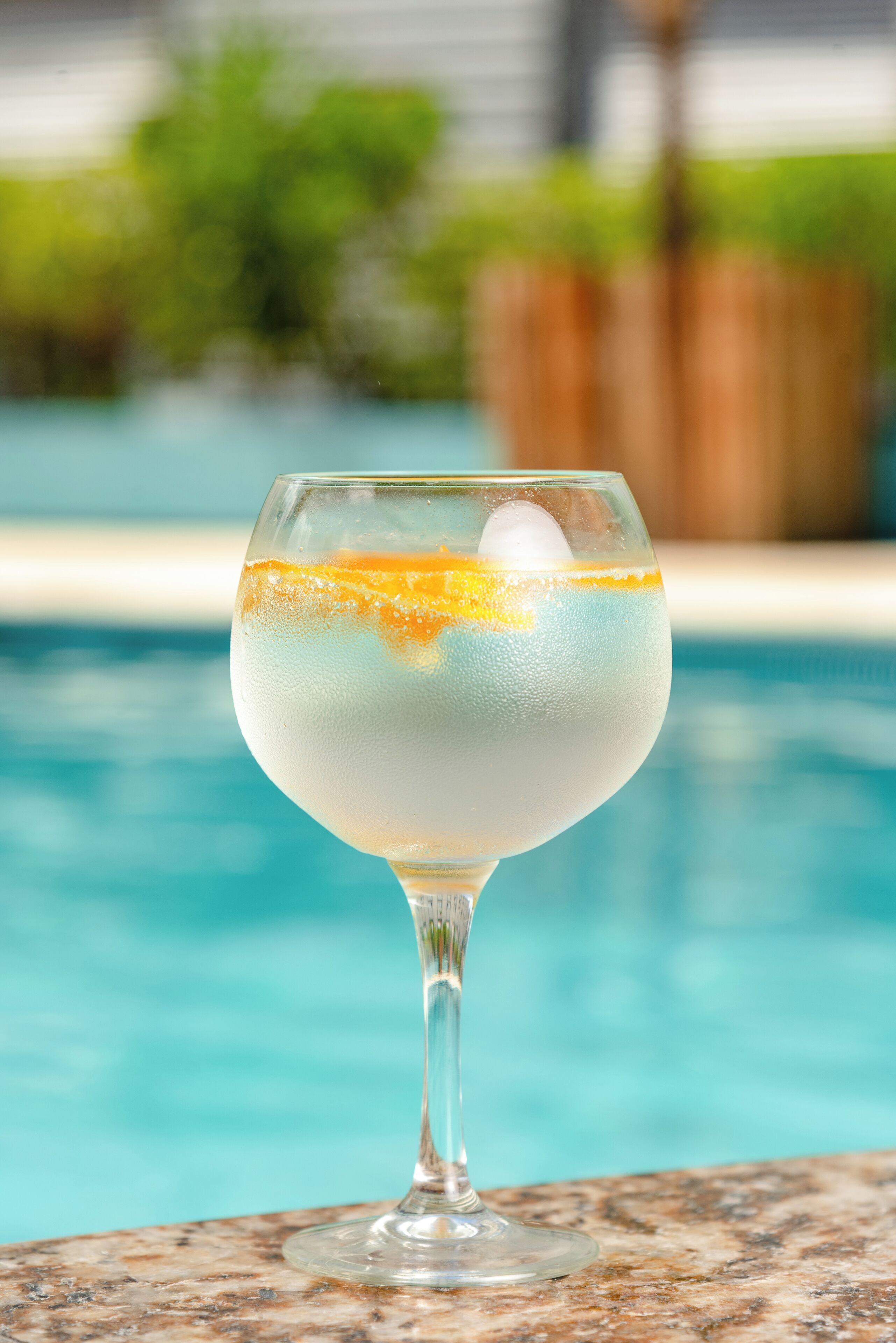 Un bicchiere da cocktail con liquido trasparente e una fetta d'arancia su una superficie in marmo, vicino a una piscina.