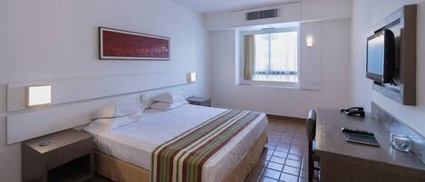 A hotel room with a bed, bedside tables, a desk, a TV, and a window.