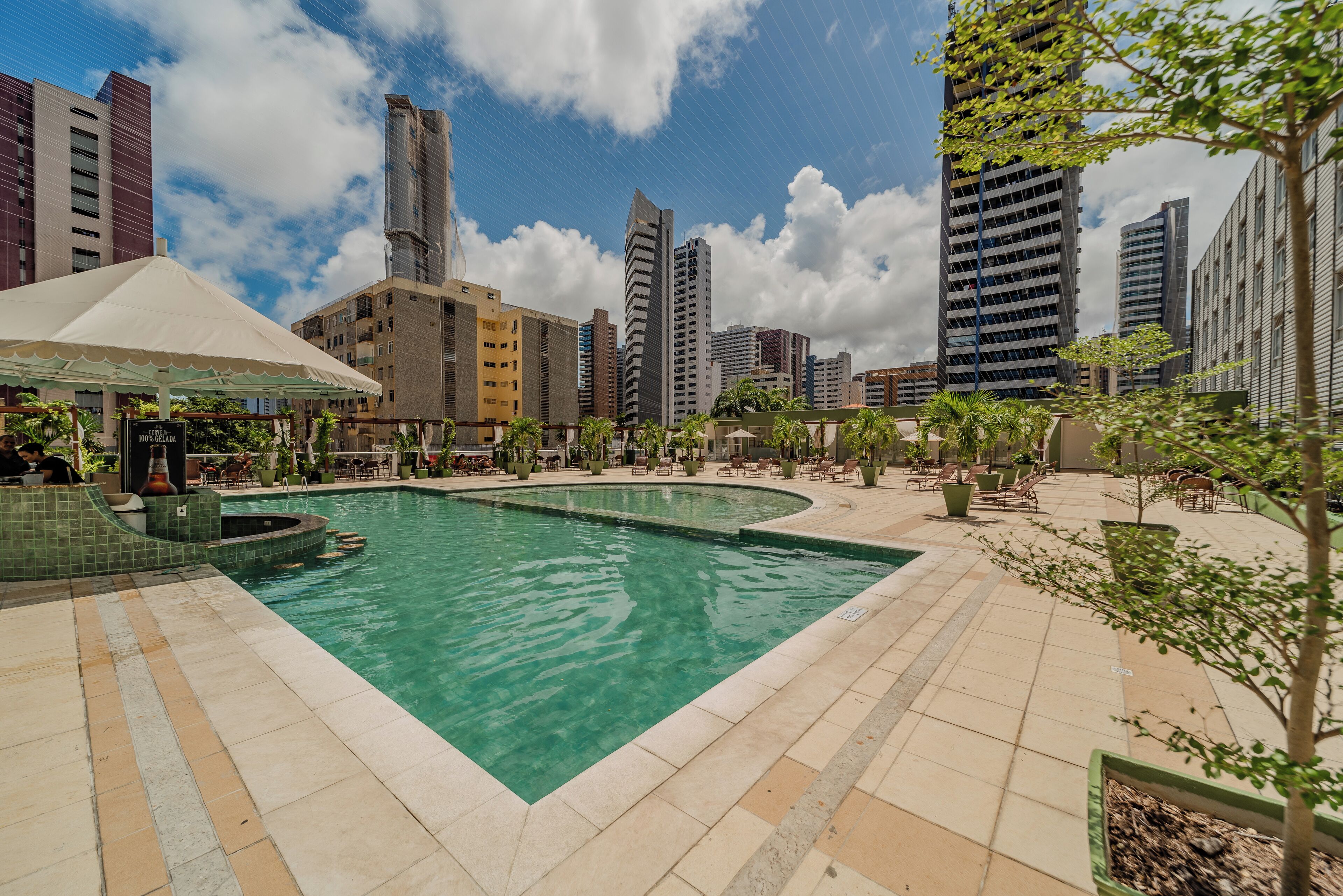 Área de piscina com deck revestido, cercada por edifícios altos e vegetação.