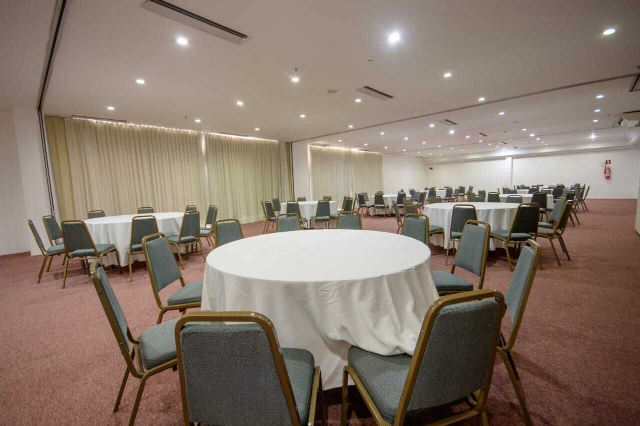 Uma sala de conferência com mesas e cadeiras redondas, piso coberto de carpete e cortinas nas janelas.
