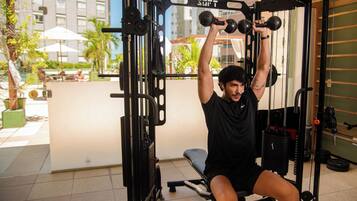 A man exercising on a Smith machine with dumbbells, located in a gym with a view of a building and palm trees outside.
