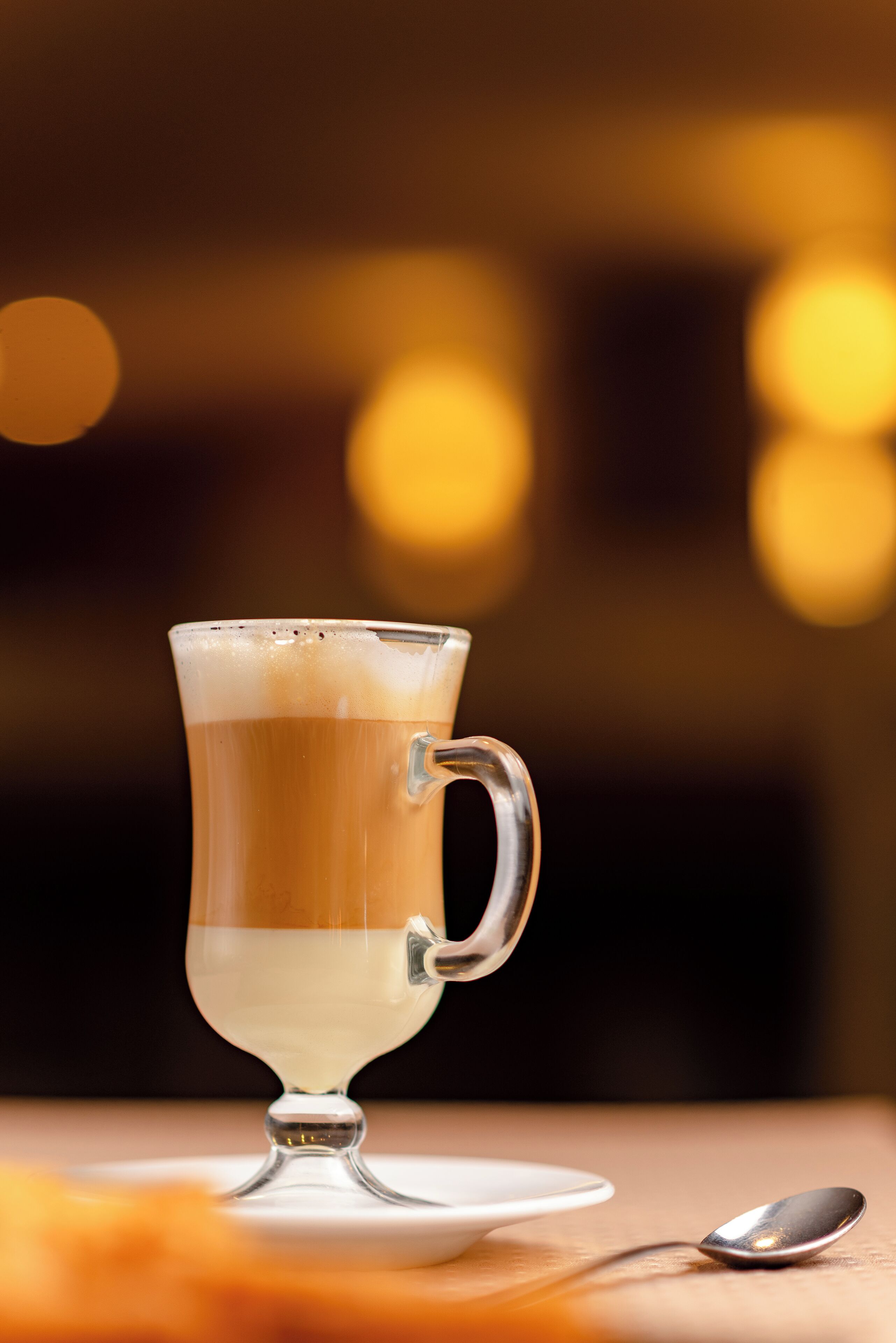 Una taza de vidrio transparente con asa, sobre un platito blanco, con una cuchara al lado.
