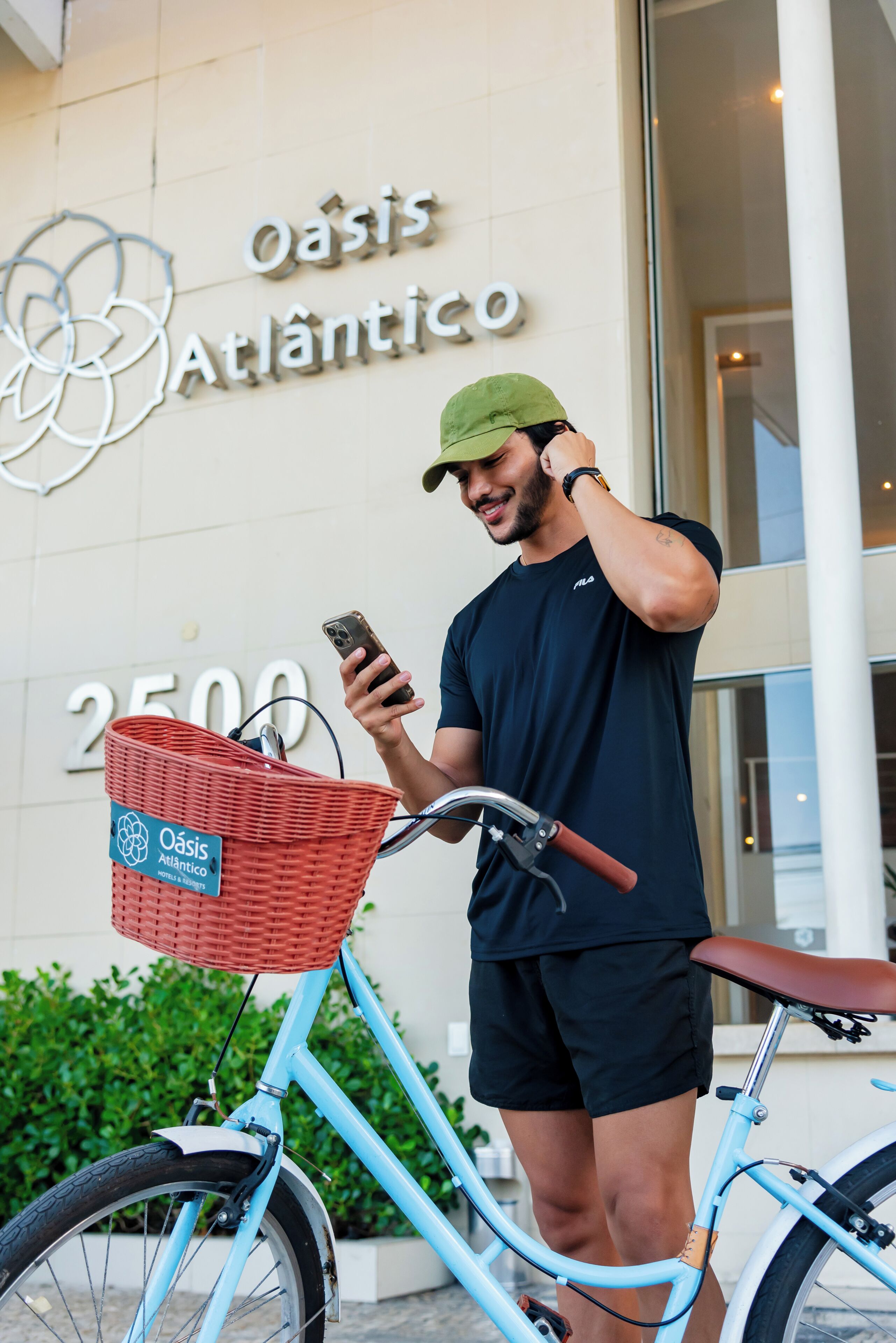 Una persona de pie junto a una bicicleta azul claro con una cesta roja, frente a un edificio con un letrero que dice 'oasis Atlanticico' y el número '2500'.