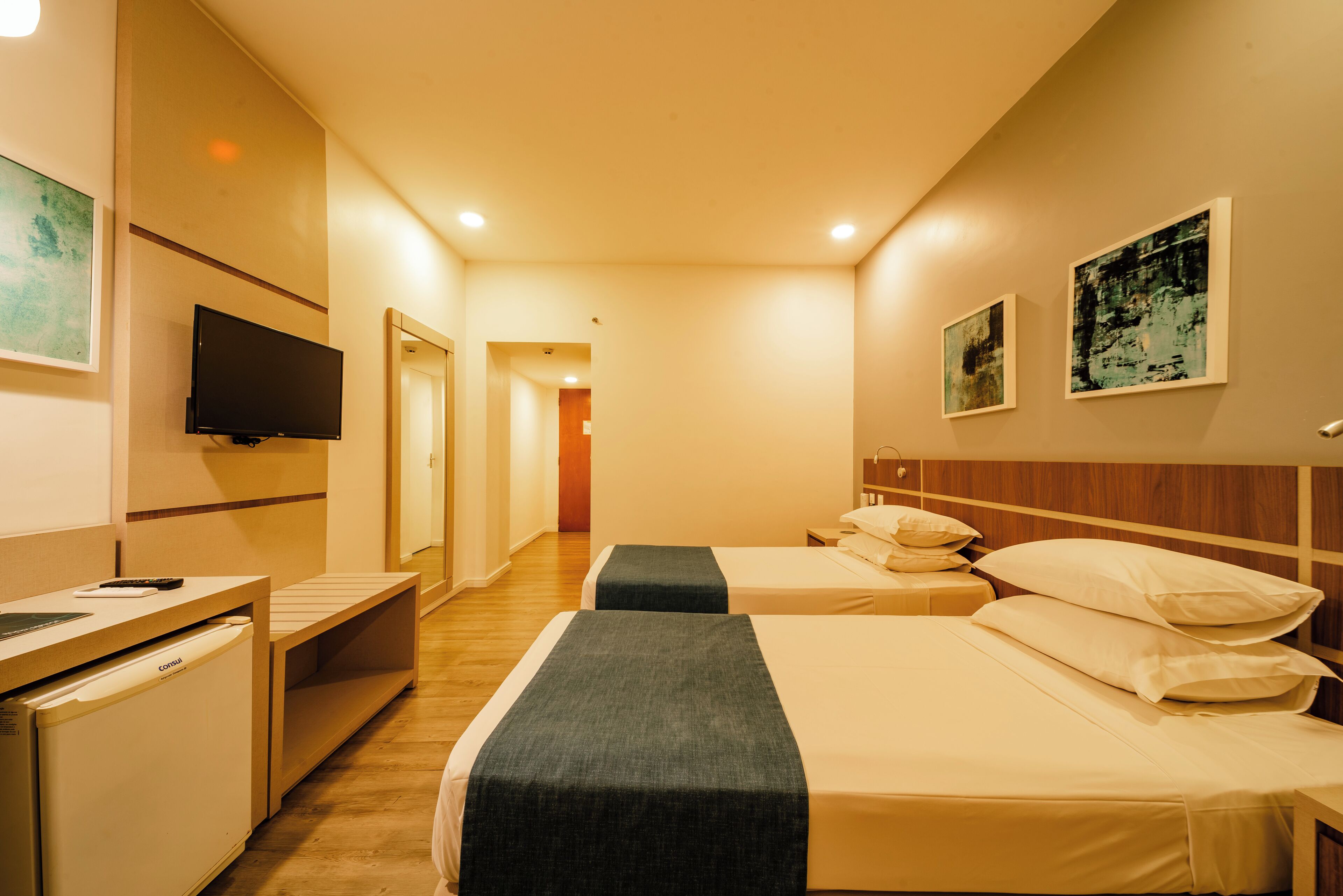 Ein Hotelzimmer mit zwei Betten, einem Fernseher, einem kleinen Schreibtisch und Bildern an den Wänden.