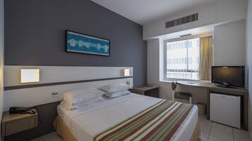 A hotel room with a bed, bedside tables, a desk with a computer, a television, and a window with curtains.