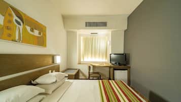 Quarto de hotel com cama, escrivaninha com computador, uma cadeira e janela com cortinas.