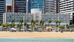 Un hotel frente a la playa con varios pisos, palmeras y sombrillas.