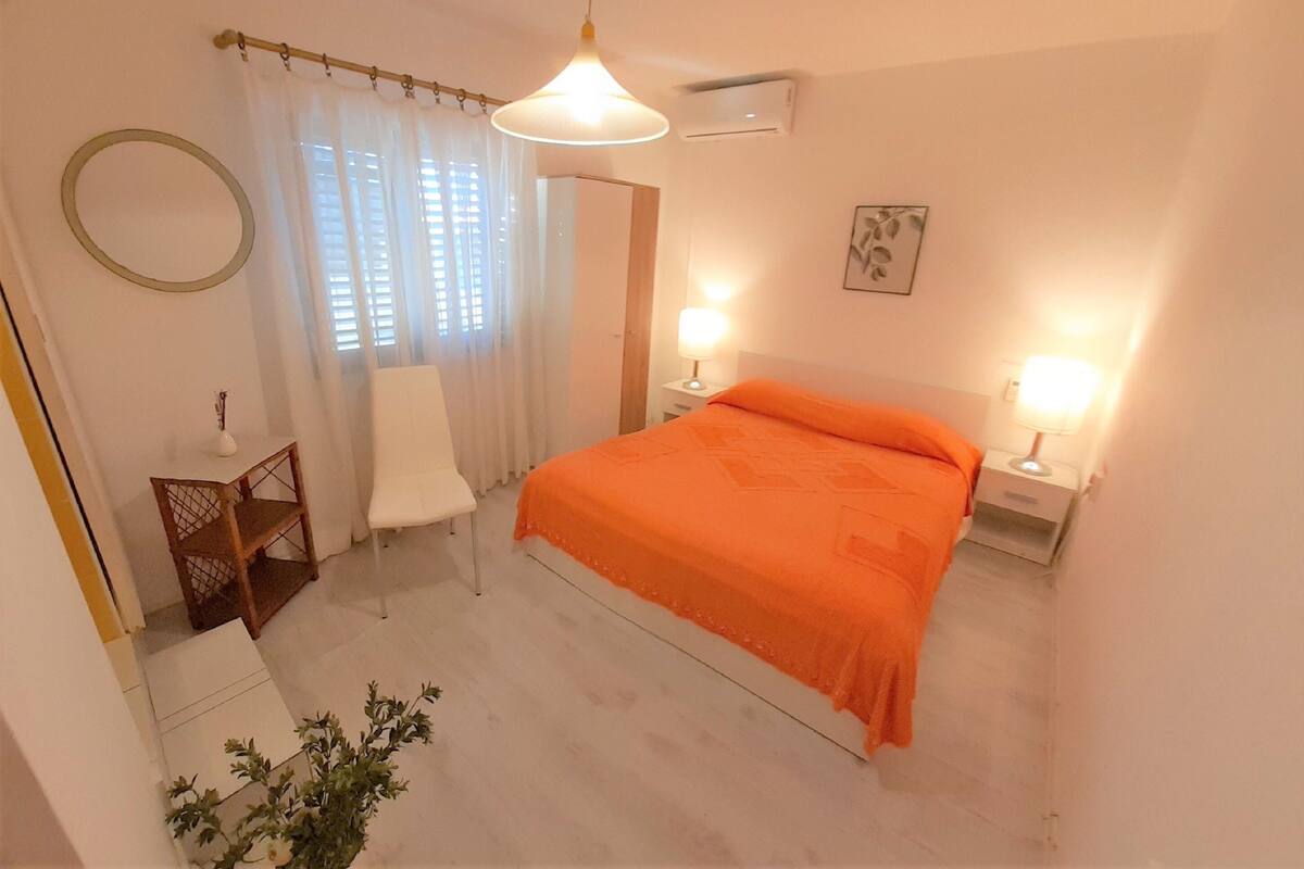 Ein Schlafzimmer mit einem Bett, zwei Nachttischlampen, einem Fenster mit Vorhängen, einem Spiegel, einem kleinen Holznachttisch und einer Topfpflanze.