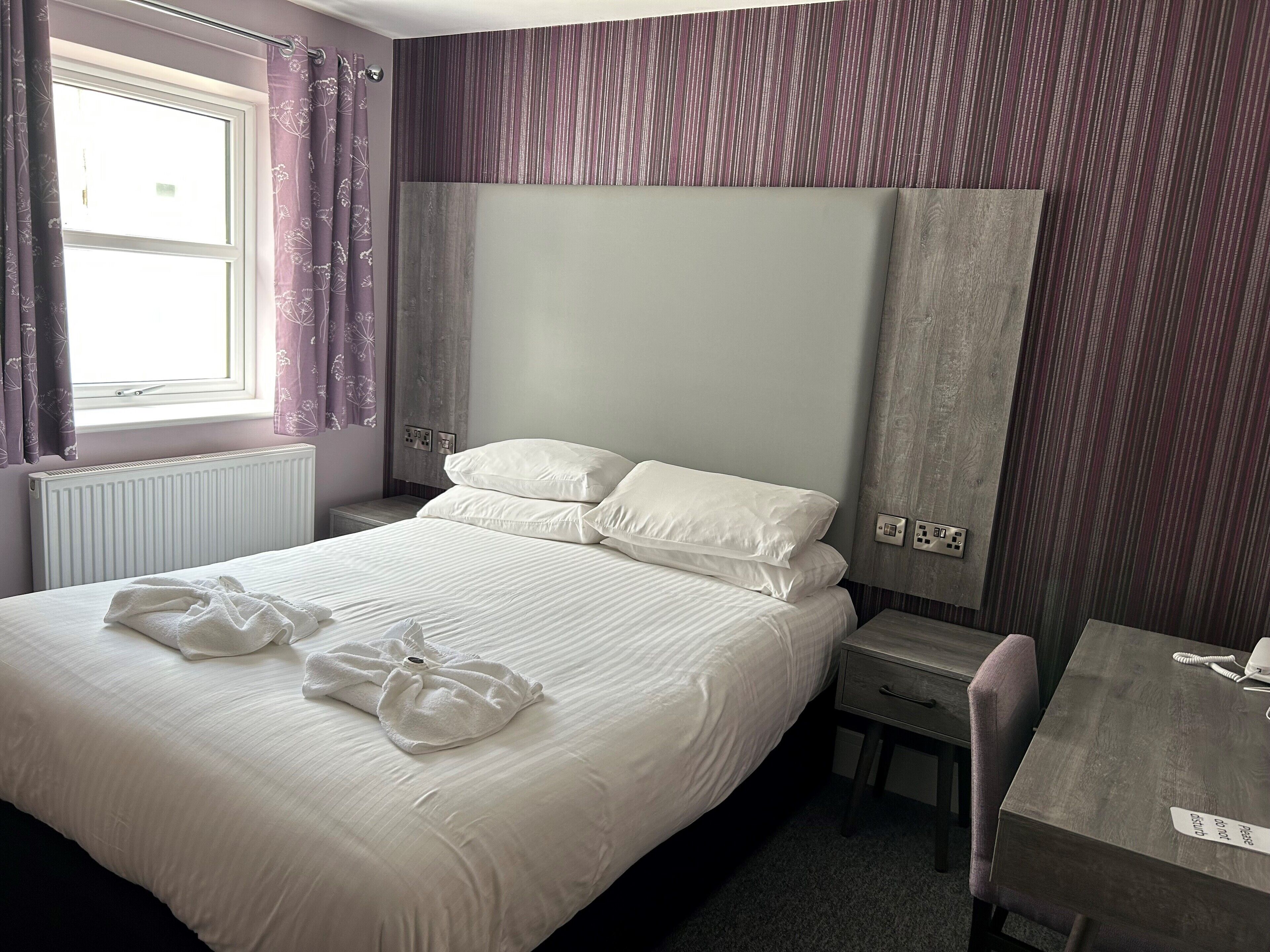 A neatly made bed with white linens and two towels, a wooden nightstand with a lamp, a small table with a telephone, and a window with purple curtains.