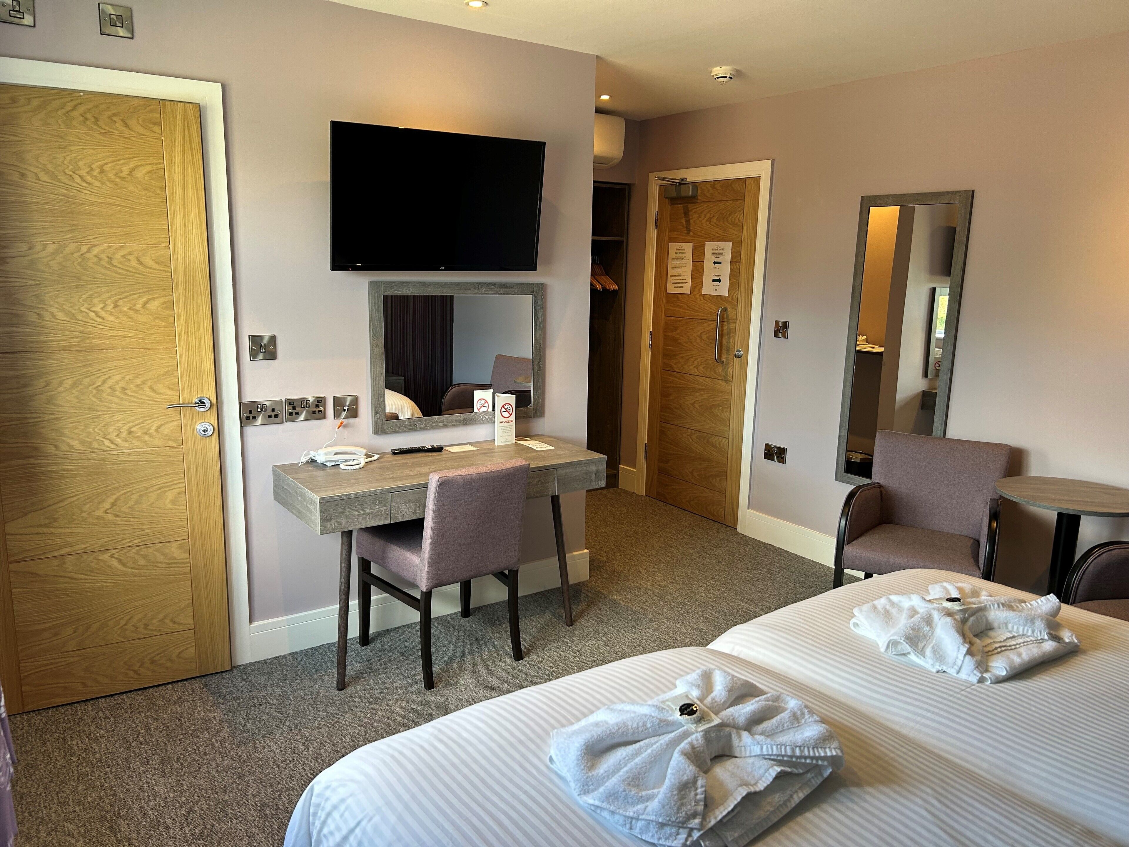 A hotel room with a wooden door, a desk with a chair, a bed with towels, a TV, and a mirror.