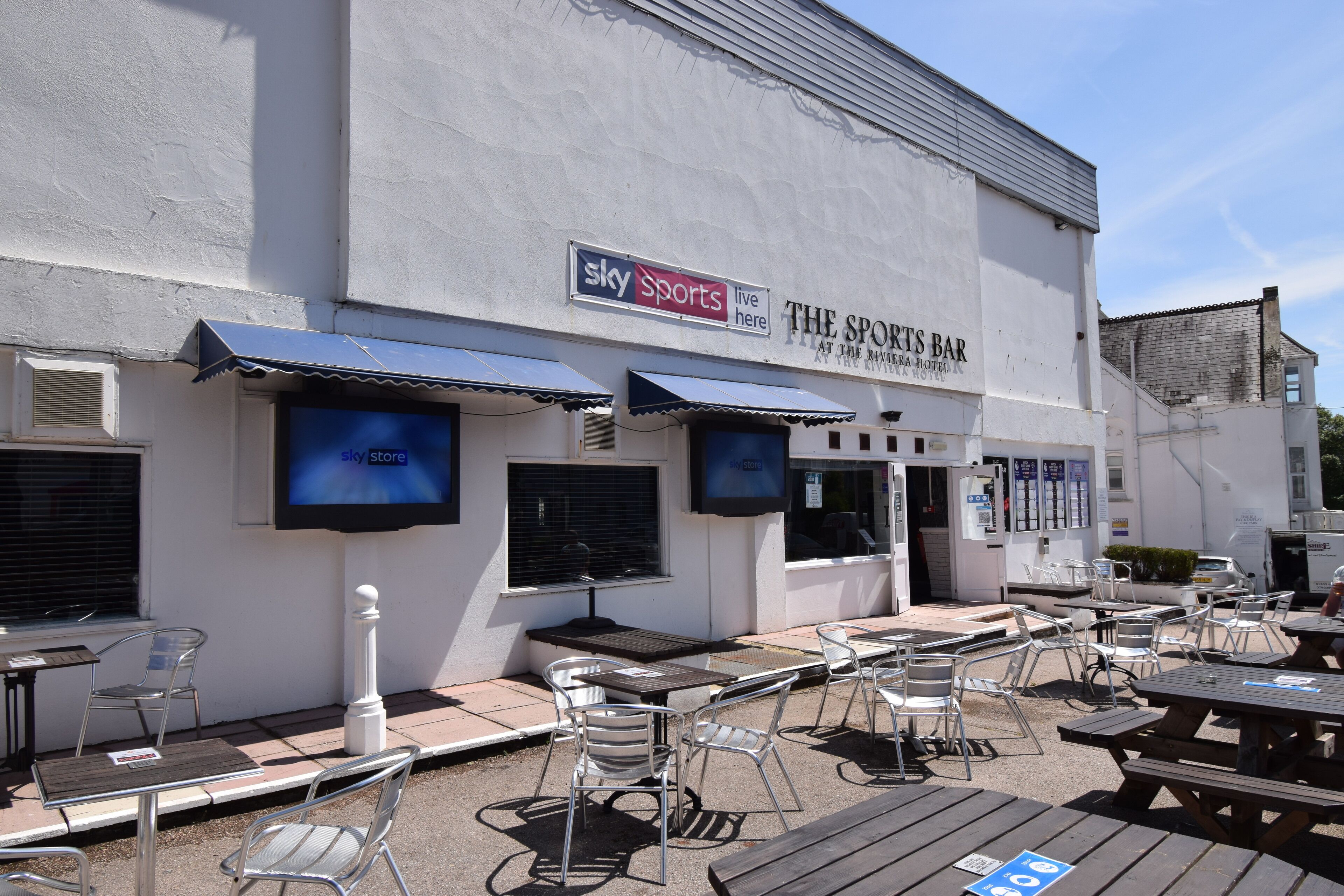 An outdoor seating area with tables and chairs, a sign for 'Sky Sports Live Here', and a building with a sign for 'The Sports Bar'.
