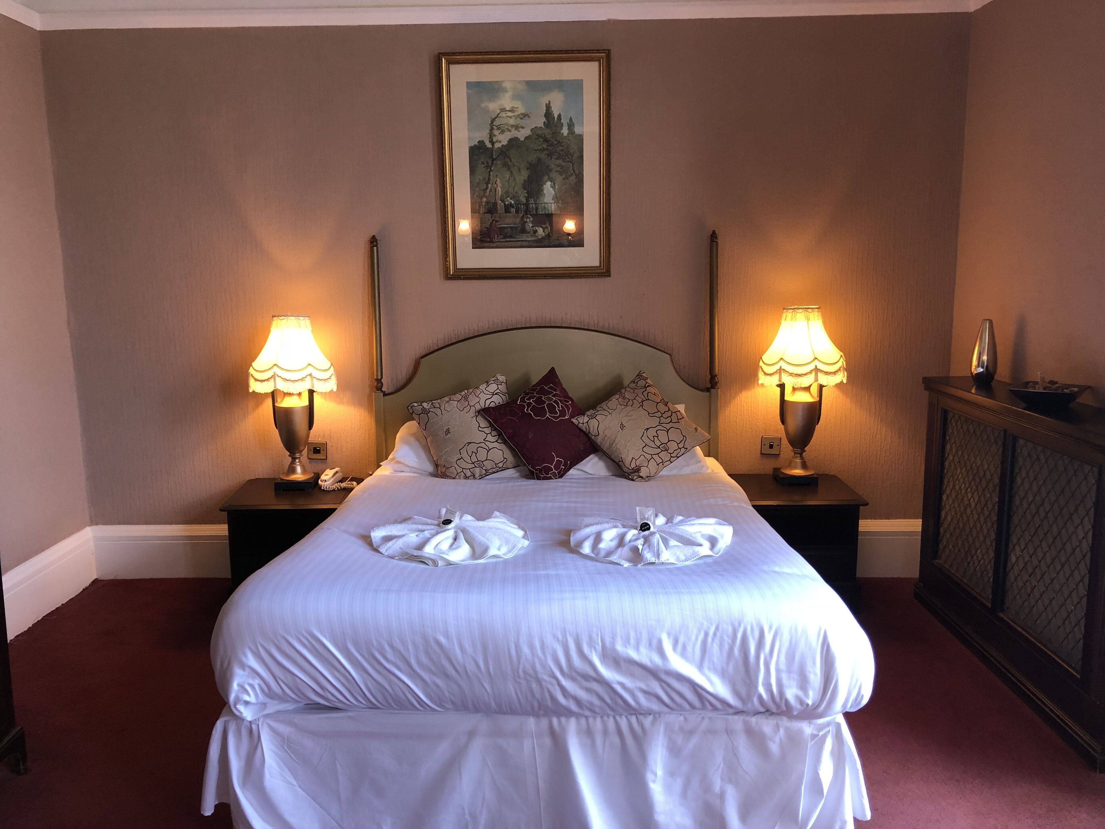 A neatly made bed with white linens, two bedside lamps, and a framed painting above the headboard.
