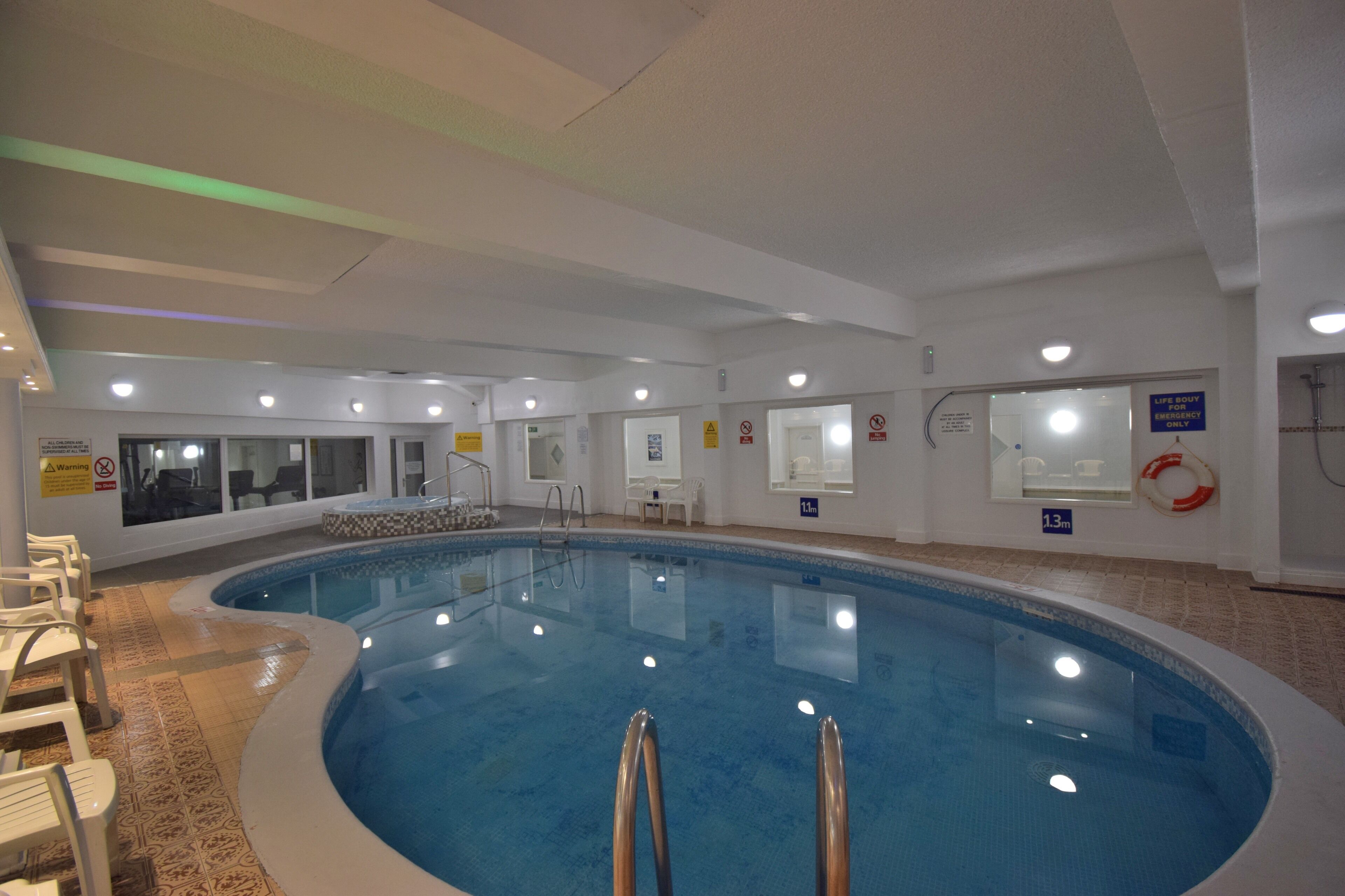Ein beheiztes Hallenbad mit klarem Wasser, umgeben von weißen Wänden und einem gefliesten Boden. An der Wand stehen Stühle und ein Rettungsring.