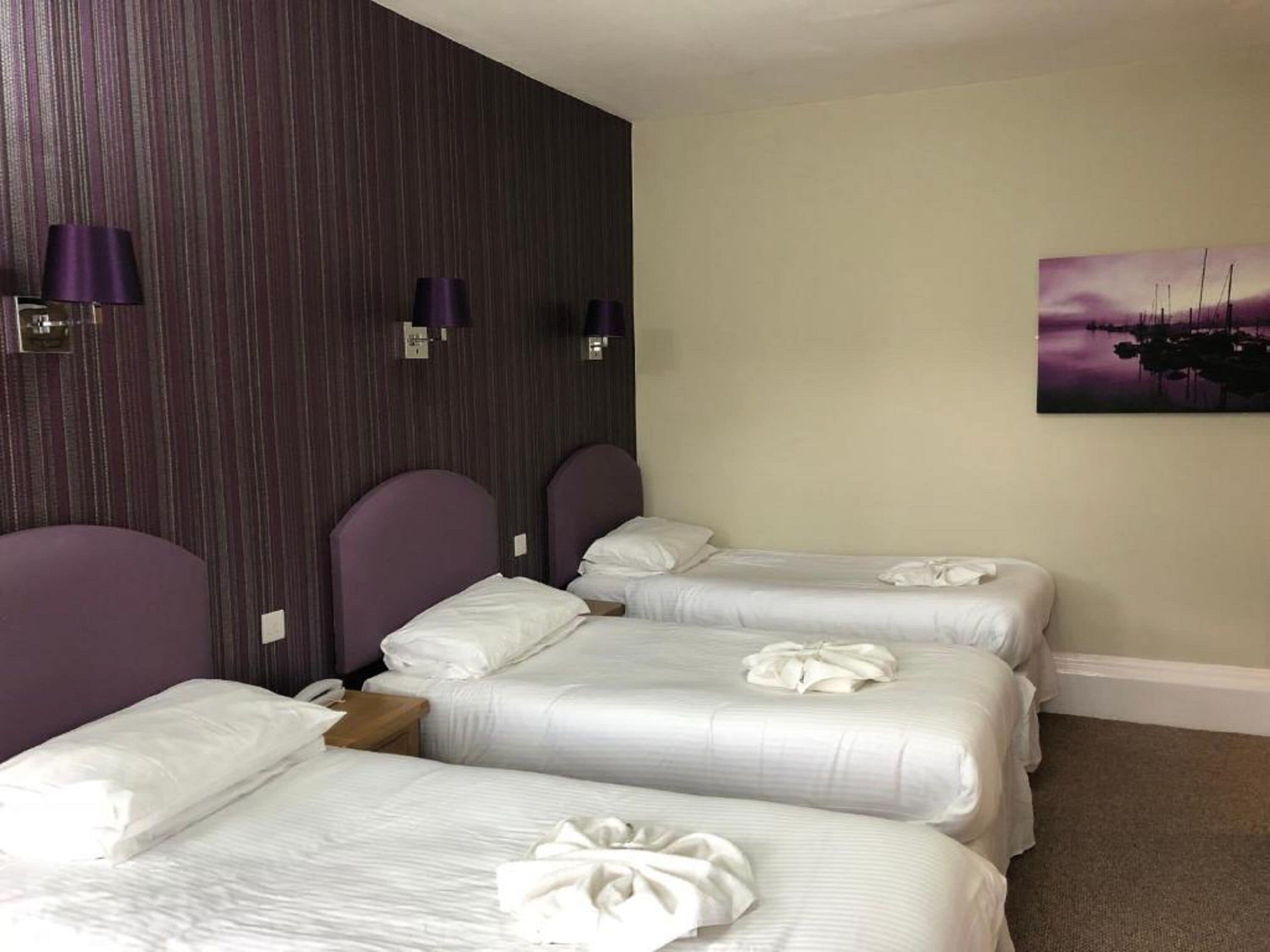 A hotel room with three beds, each with white linens and a towel on the bed. There are purple headboards and wall-mounted lamps. A painting of a landscape is on the wall.
