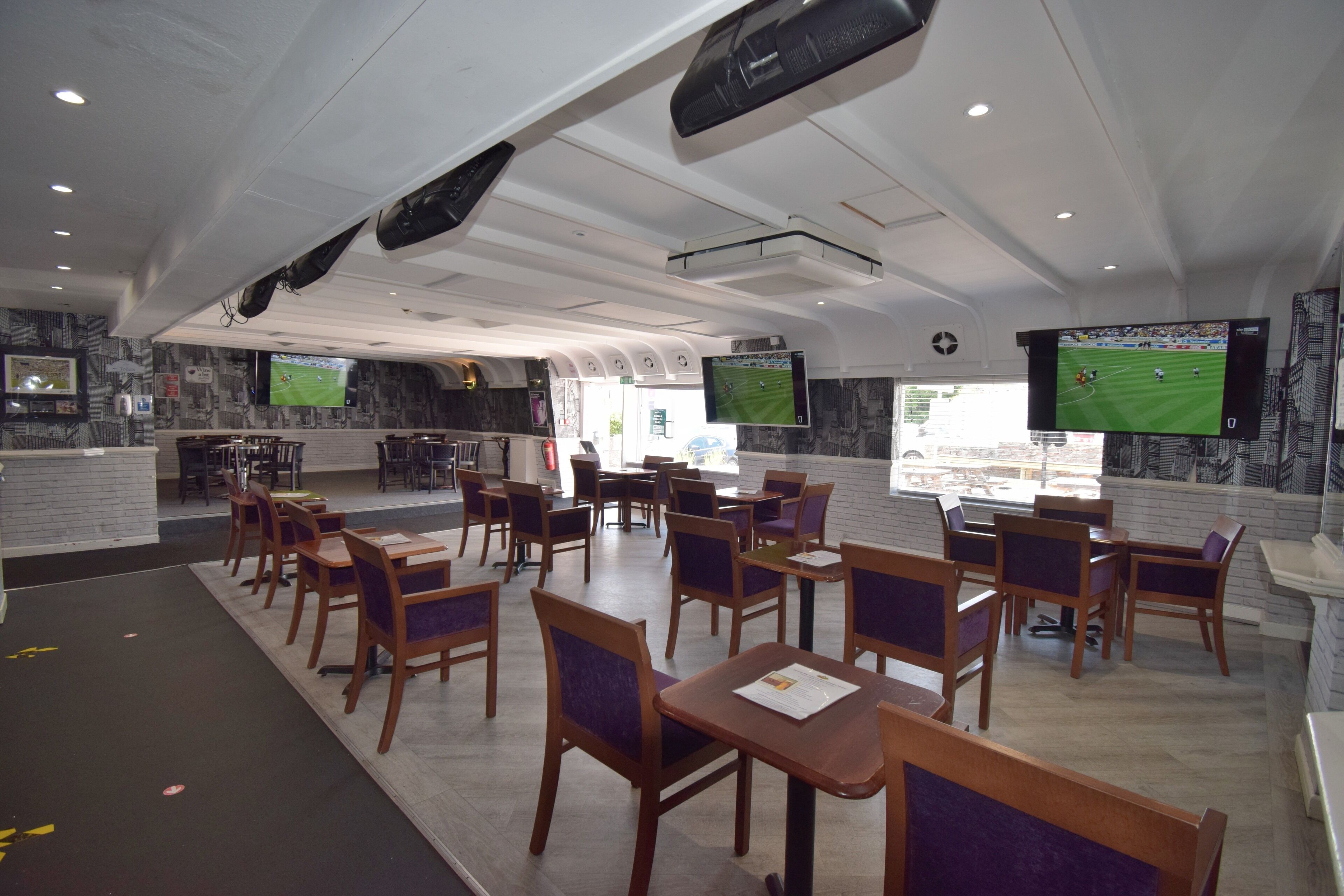 Un vaste espace intérieur aménagé avec différents types de sièges, notamment des tables et des chaises, ainsi que deux grands écrans diffusant des événements sportifs.