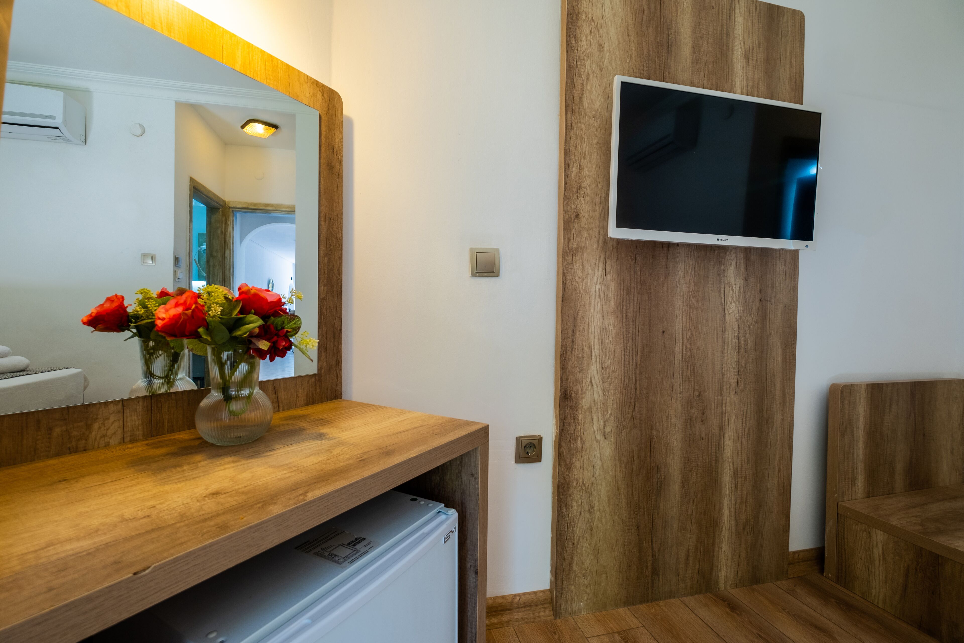A modern bathroom with a wooden vanity, a mirror, a flat-screen TV mounted on the wall, and a refrigerator under the vanity.
