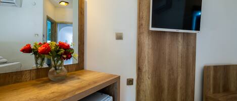 A modern bathroom with a wooden vanity, a mirror, a flat-screen TV mounted on the wall, and a refrigerator under the vanity.