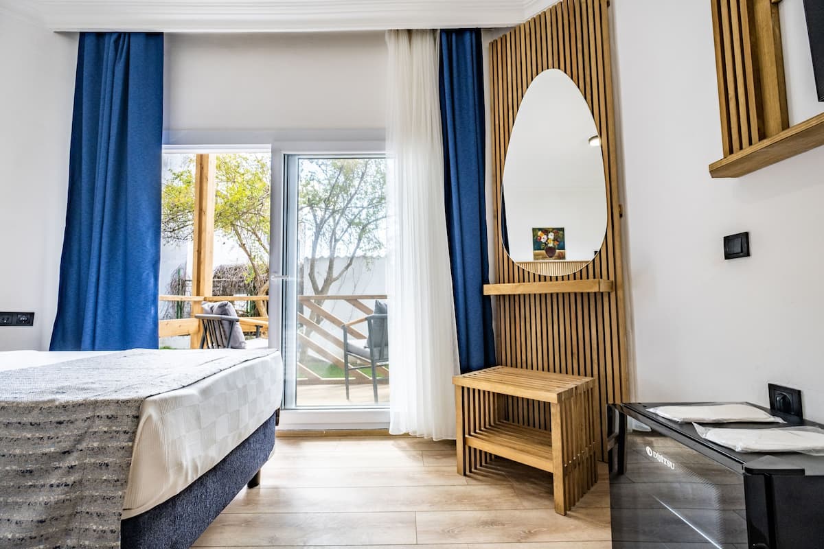 A modern hotel room with a bed, a wooden bench, a mirror, and a view of the outdoors.