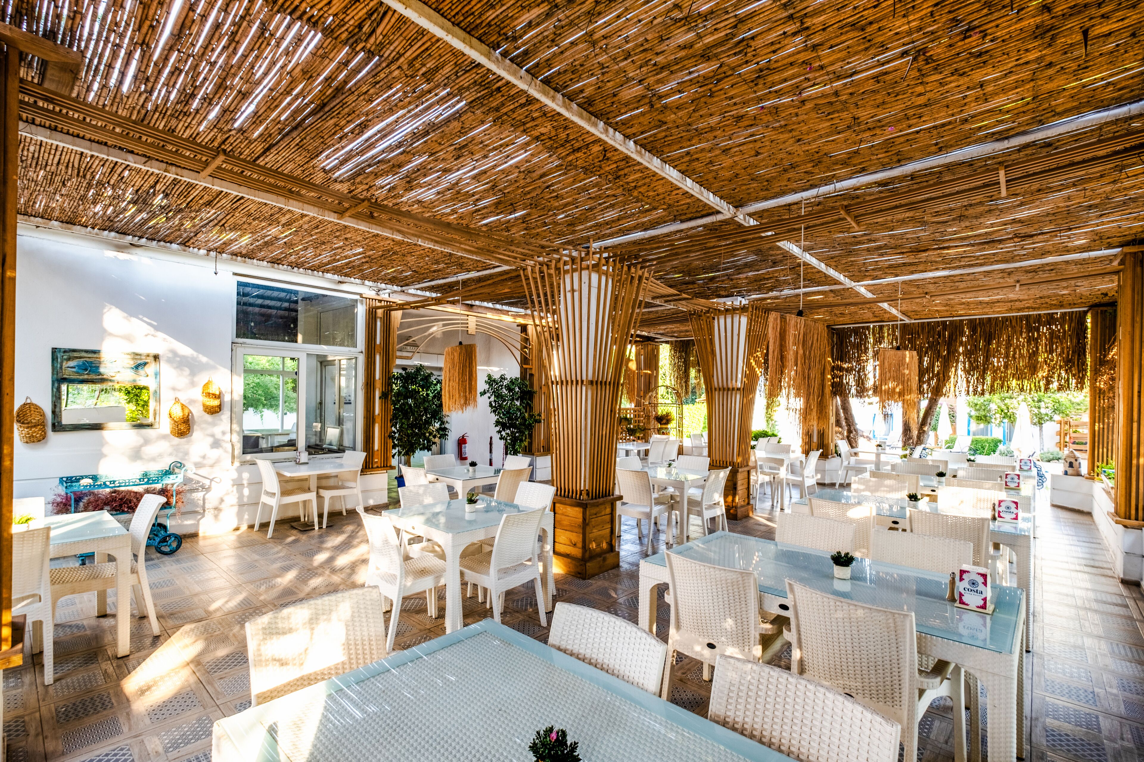 An outdoor dining area with white wicker furniture, a bamboo ceiling, and large windows allowing natural light.