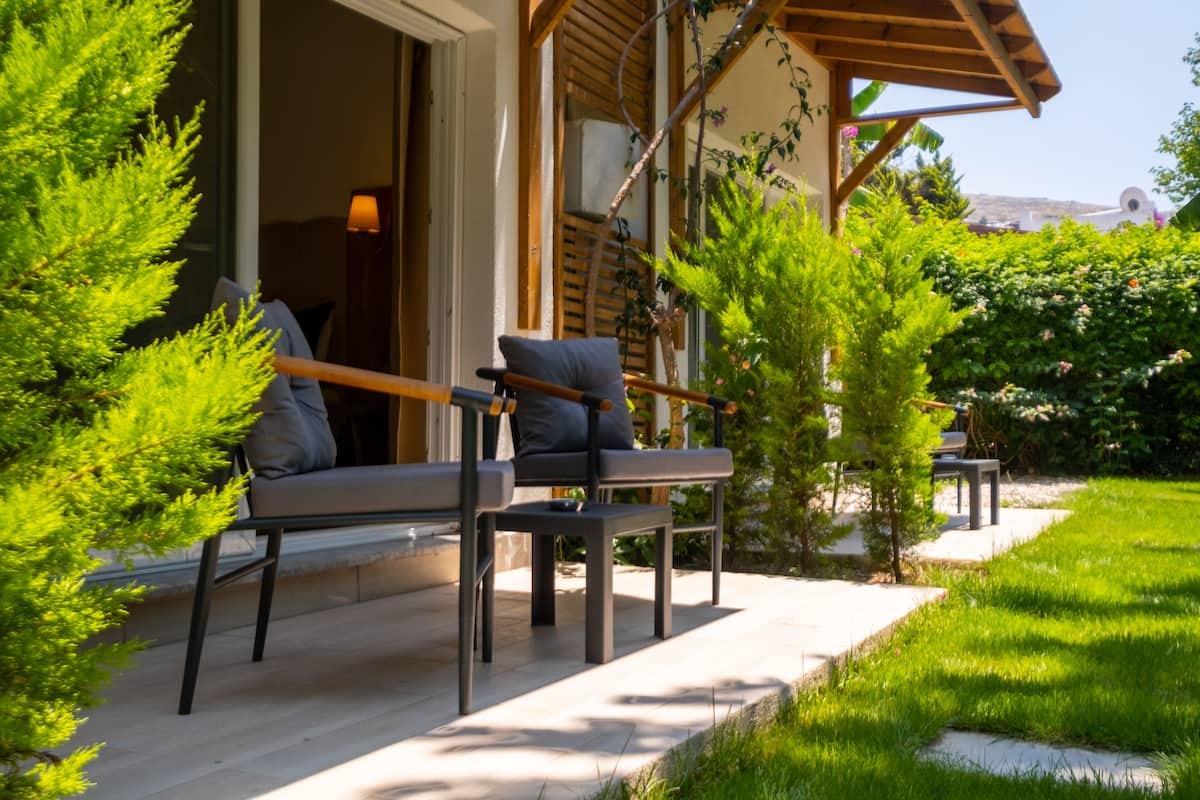 An outdoor seating area with two chairs and a table, surrounded by greenery and a building with a wooden pergola.