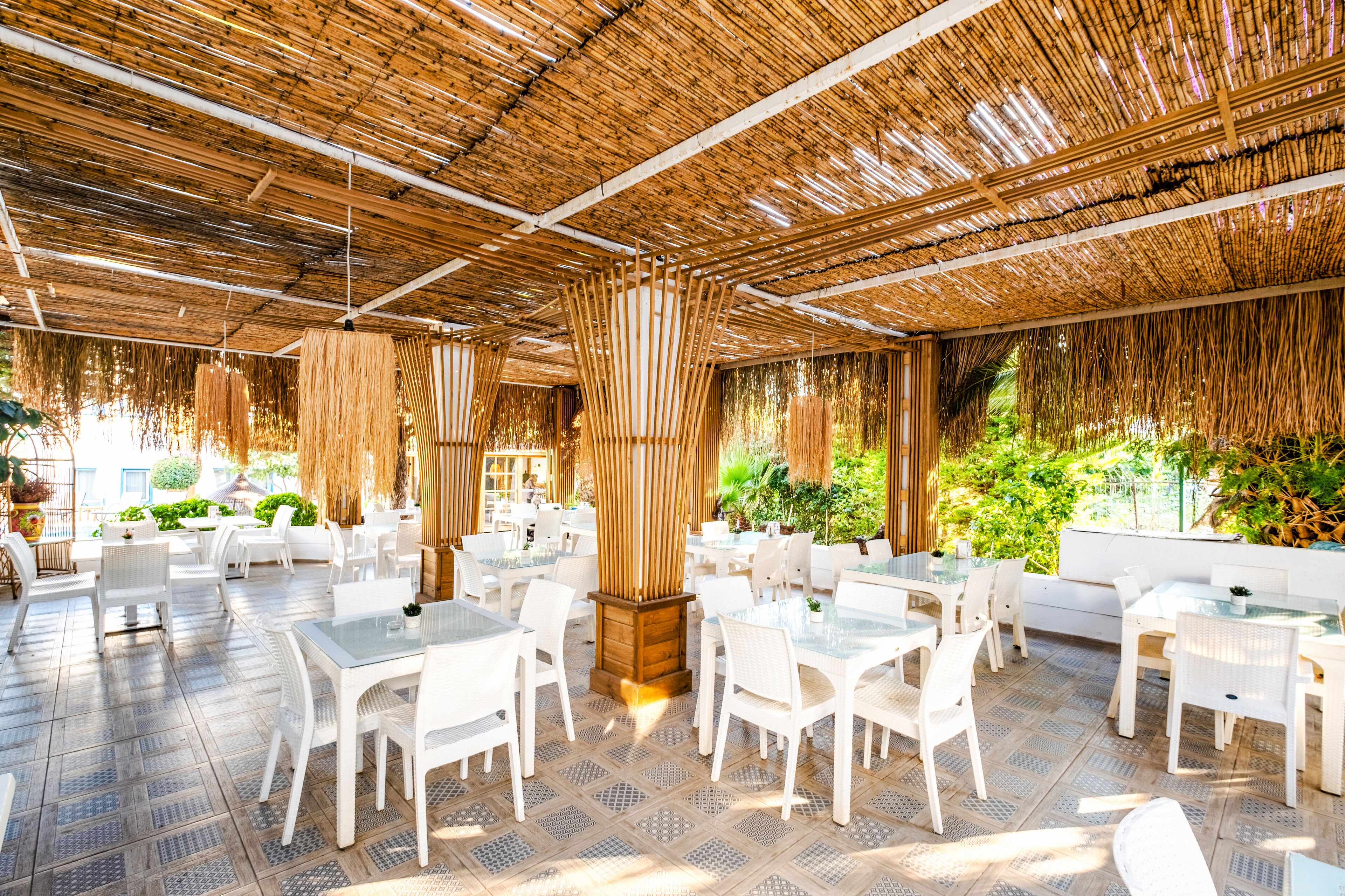 Un área de comedor al aire libre con mesas y sillas blancas, techo de paja y follaje de fondo.