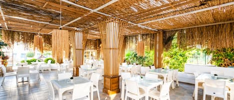 Un área de comedor al aire libre con mesas y sillas blancas, techo de paja y follaje de fondo.