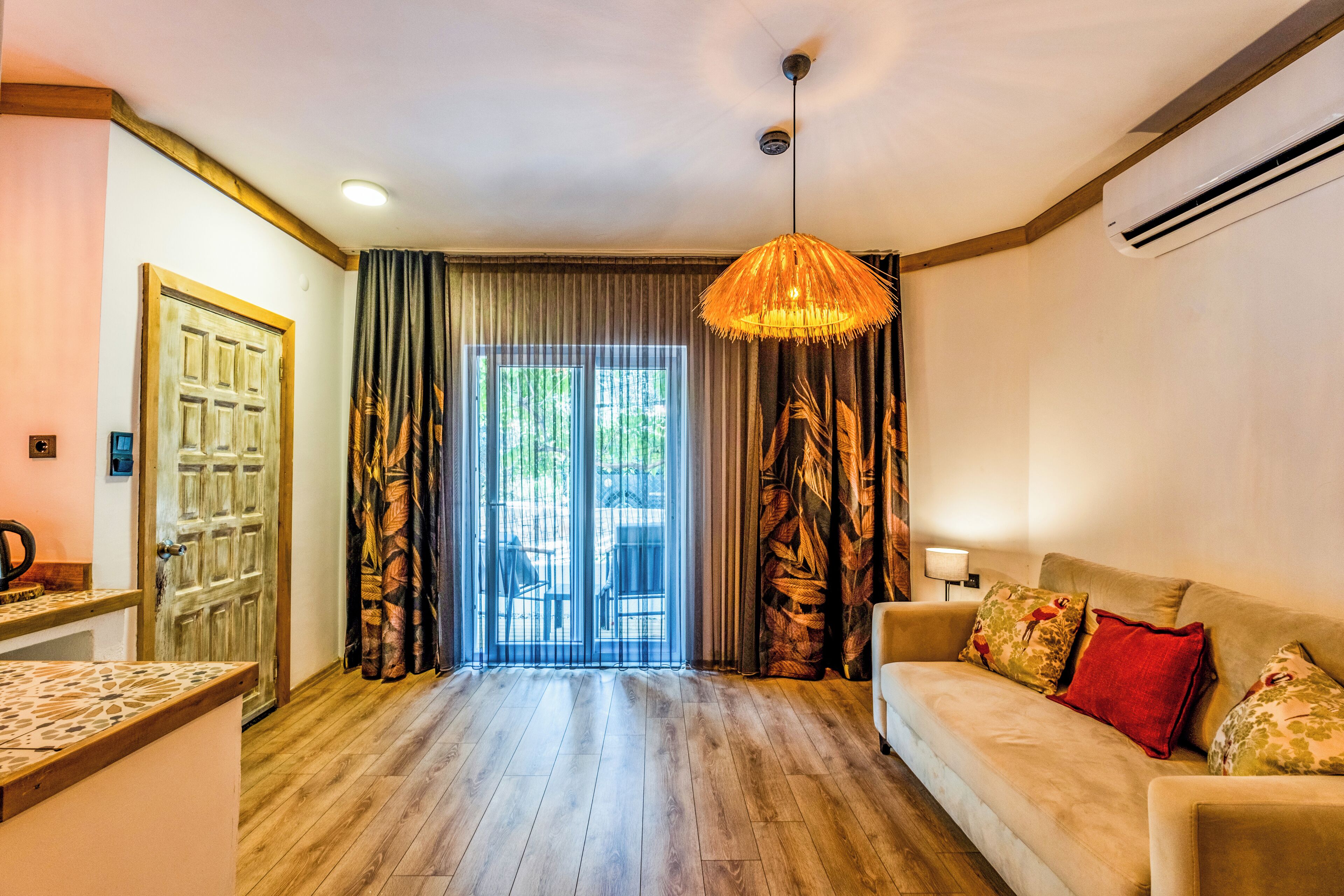 Amplia sala de estar con piso de madera, sofá color beige con cojines estampados, lámpara colgante y puerta corrediza de vidrio que da a un área exterior.