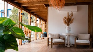 A room with wooden flooring, a white table with a vase of pampas grass, two white cushioned chairs, and large windows allowing natural light. - Costa Bodrum City (Bodrum)